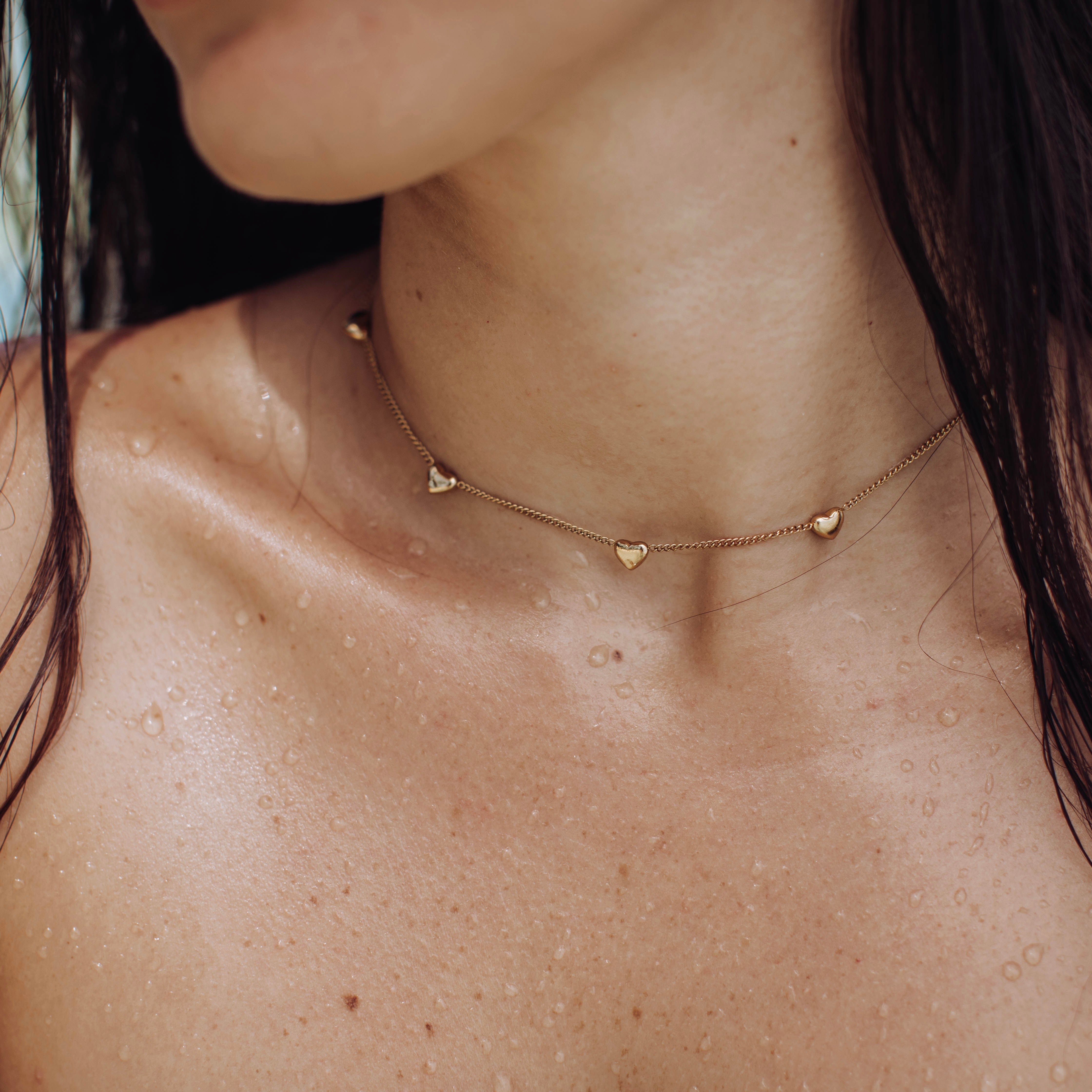 A close-up of a woman’s neck and shoulders with wet skin showcases the Waterproof Paige Necklace, a delicate gold plated stainless steel piece with small heart-shaped charms. Her dark hair falls partially over her shoulder.